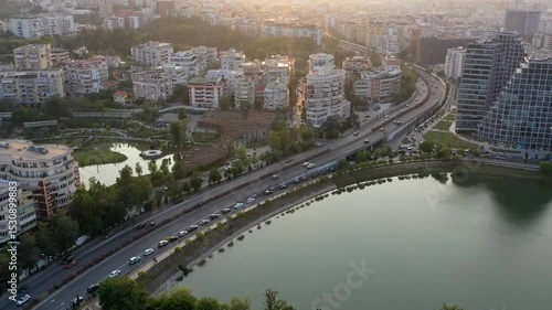 Forward drone hyperlapse over Tirana’s Grand Lake, capturing fast-moving traffic and the architectural contrast between modern towers and residential blocks at golden hour.