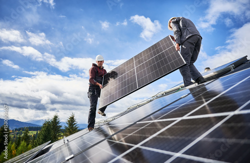 Roofers building solar panel system on roof of house. Men workers in helmets carrying photovoltaic solar module outdoors. Concept of alternative and renewable energy.