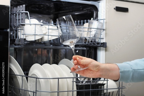 Man holding a wine glass near open dishwasher filled with clean dishes and glassware in a kitchen
