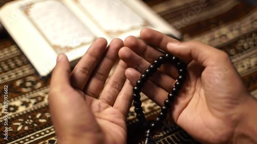 Hands Holding Prayer Beads Over a Religious Book. Close-Up of Devotion with an Open Quran. Spiritual Moment with Black Tasbih Beads. Intricate Fabric and a Book of Faith.