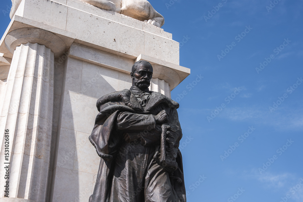 Obraz premium The Tisza Istvan Memorial. A bronze statue of a man in formal attire holding a scroll stands on a white stone pedestal with fluted columns and a sculpted lion in Budapest, Hungary.