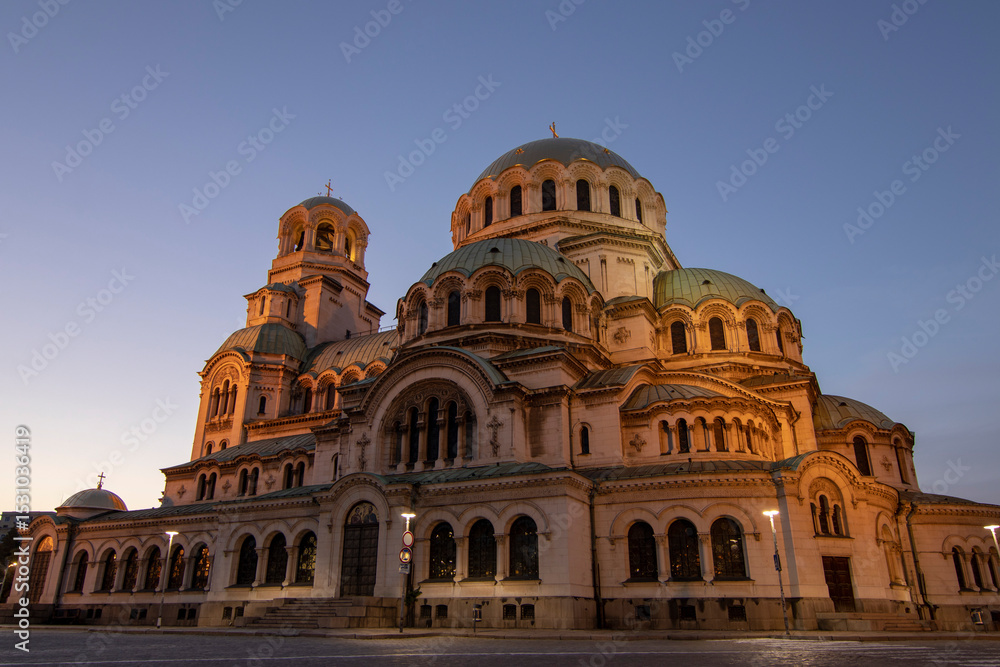 Obraz premium Alexander Nevsky Cathedral in Sofia illuminated by warm sunset light, highlighting its impressive Neo-Byzantine architecture and intricate domes against a clear evening sky.