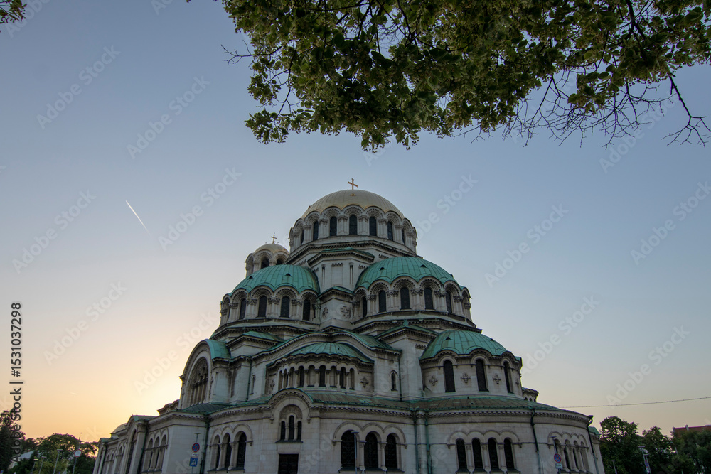 Fototapeta premium Alexander Nevsky Cathedral in Sofia framed by tree branches at sunset, showcasing its majestic Neo-Byzantine architecture and iconic green domes against a clear evening sky.