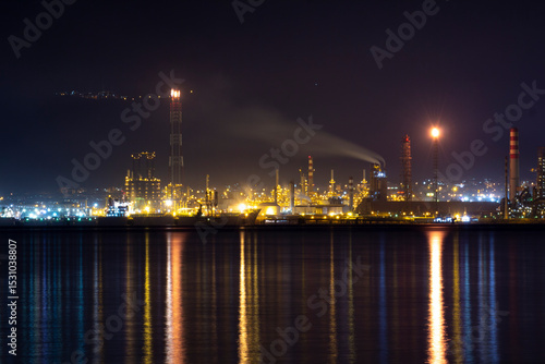 Night view of İzmit Tüpraş oil refinery with bright industrial lights reflecting on calm water, featuring chimneys emitting smoke and a vibrant urban industrial landscape at night.

