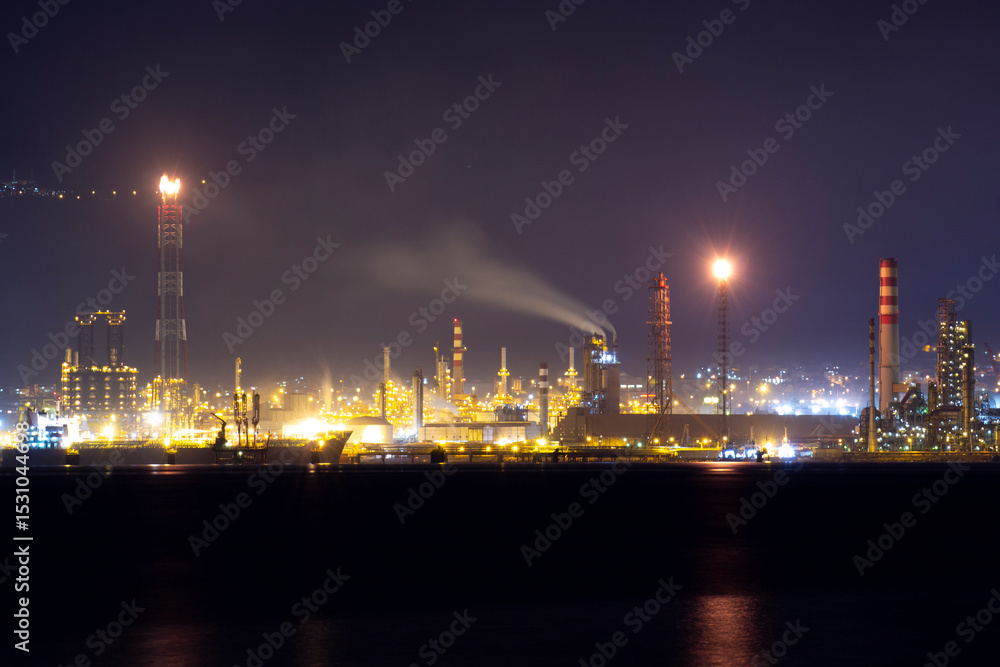 Fototapeta premium Brightly lit oil refinery at night with flare stacks, smoke emissions, and glowing chimneys by the sea, reflecting industrial energy production and environmental pollution issues.