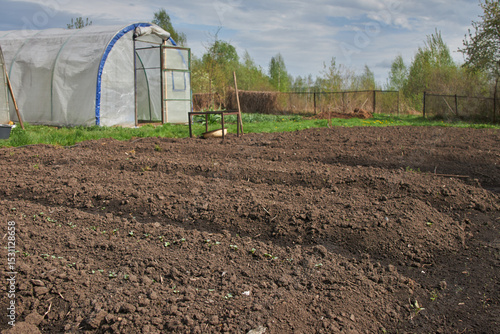 A freshly tilled garden plot awaits planting beside a greenhouse under a cloudy sky.