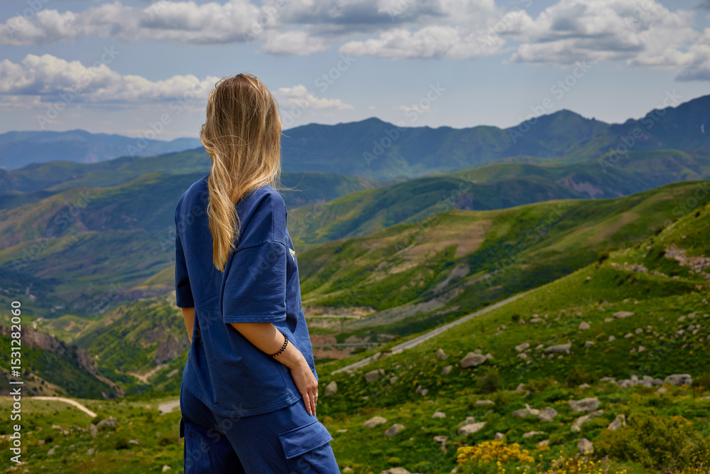 Naklejka premium Woman standing on a mountain peak with arms outstretched, enjoying a breathtaking panoramic view of green hills and valleys under a partly cloudy sky, symbolizing freedom and connection with nature.