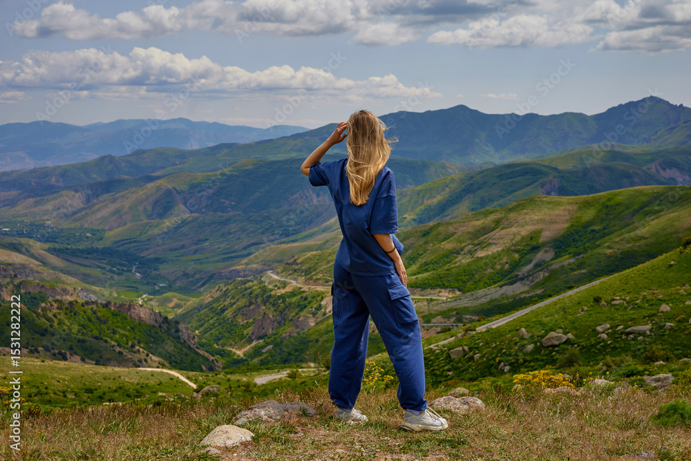 Fototapeta premium Woman standing on a mountain peak with arms outstretched, enjoying a breathtaking panoramic view of green hills and valleys under a partly cloudy sky, symbolizing freedom and connection with nature.
