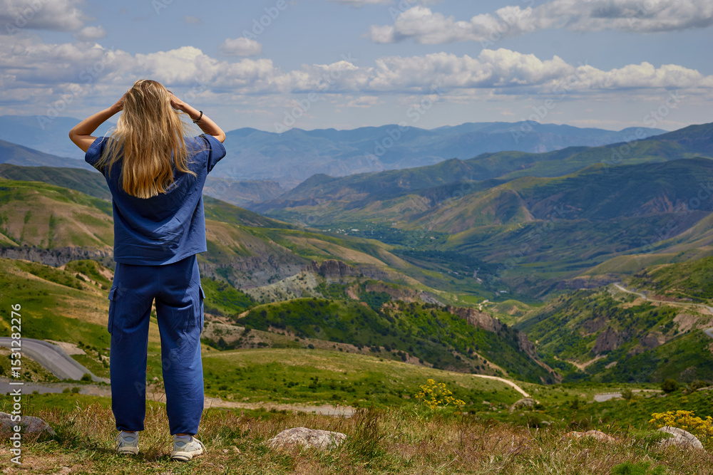 Fototapeta premium Woman standing on a mountain peak with arms outstretched, enjoying a breathtaking panoramic view of green hills and valleys under a partly cloudy sky, symbolizing freedom and connection with nature.