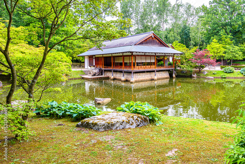 Oriental wooden house next to pond surrounded by lush green foliage trees, calm waters with reflection on surface, Japanese garden on cloudy spring day, Hasselt, Belgium