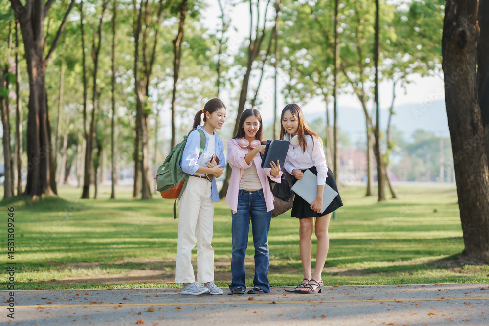 Three young women, seemingly college students, are standing in a park, looking at a digital tablet and discussing Three young women, seemingly college students, are standing in a park, looking at a digital tablet and discussing