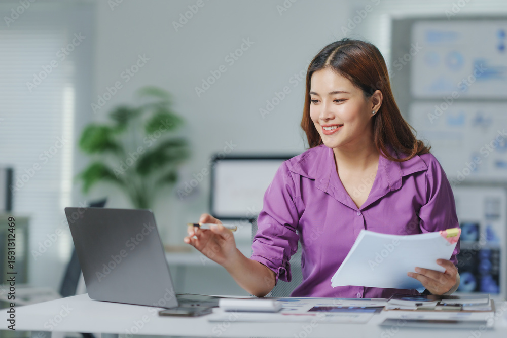 Young Asian businesswoman working at a modern office desk, using a laptop and holding documents while engaging in a video call Young Asian businesswoman working at a modern office desk, using a laptop and holding documents while engaging in a video call