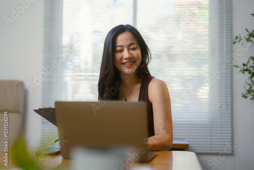 Smiling young Asian businesswoman working from home using laptop and taking notes on clipboard Free Download Smiling young Asian businesswoman working from home using laptop and taking notes on clipboard - GFXLoad