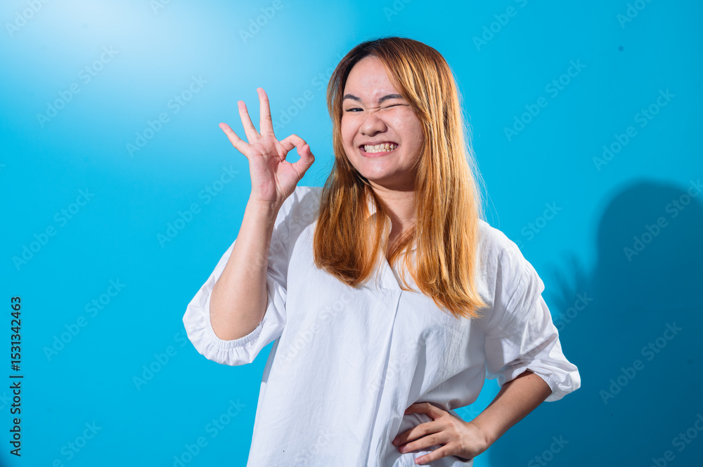 Fototapeta premium Asian woman with long straight hair in a white shirt smiles brightly while making an OK hand gesture with one hand and winking playfully against a plain blue background in studio lighting