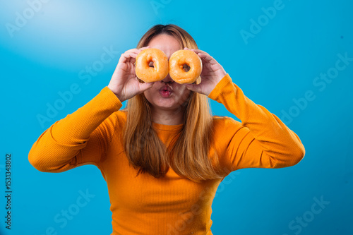 Wallpaper Mural Asian woman playfully holding two glazed donuts over her eyes like glasses, laughing with mouth open, wearing orange top on vibrant blue background Torontodigital.ca