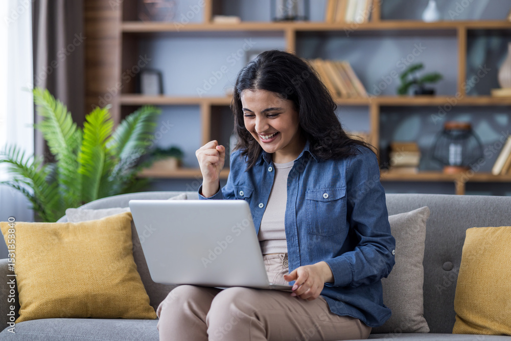 Fototapeta premium Happy Indian young woman sitting on the sofa at home, holding a laptop on her lap and rejoicing in success, showing a victory gesture