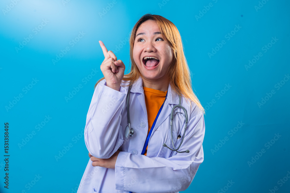 Excited Asian female doctor in white coat and orange shirt pointing upward with wide open mouth and enthusiastic expression, standing against bright blue background in studio setting Excited Asian female doctor in white coat and orange shirt pointing upward with wide open mouth and enthusiastic expression, standing against bright blue background in studio setting