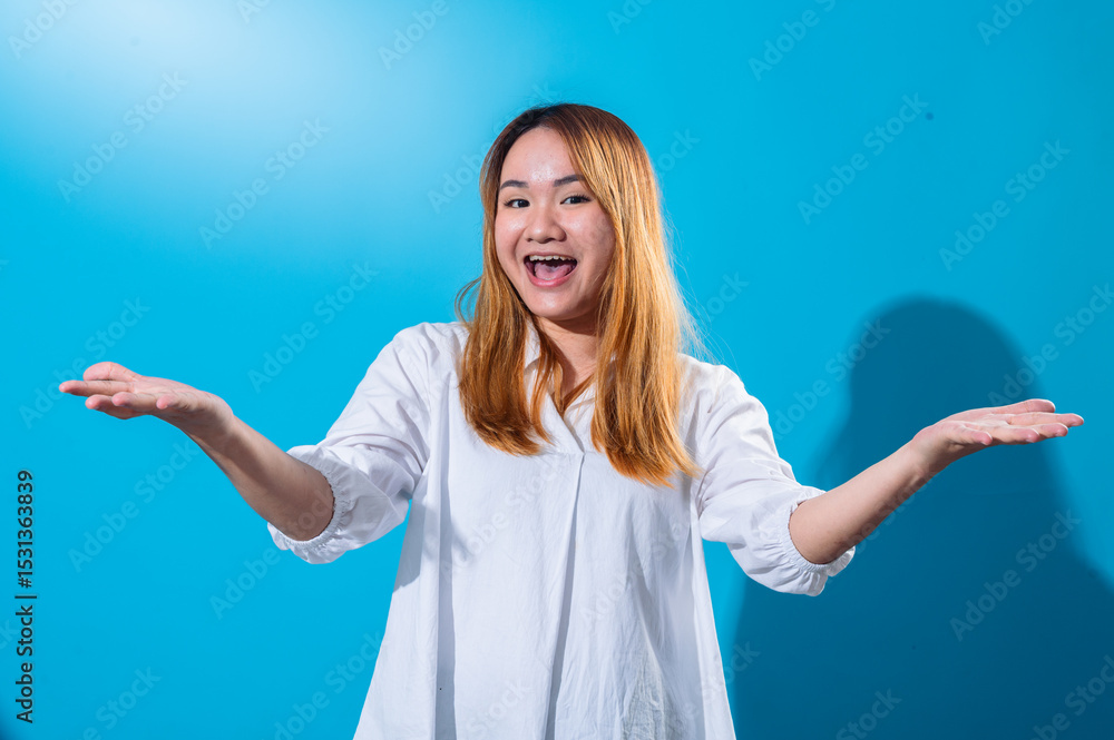 Fototapeta premium Asian woman with long straight hair wearing white shirt smiles widely while holding both hands open to the sides in welcoming gesture, standing against a bright blue background in studio light