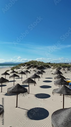 Drone approaching a straw parasol on the sandy beach of Caparica near Lisbon, Portugal. Blue sky, warm sunlight, relaxed atmosphere and gentle cinematic motion.

