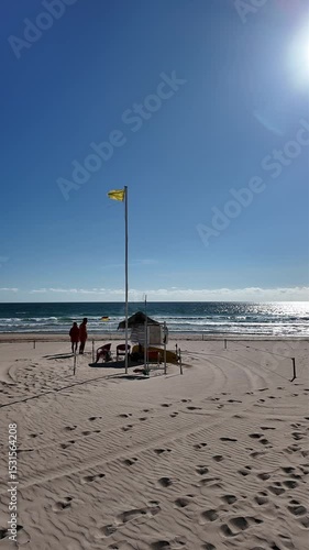 Drone alternating between straw parasols and red beach safeguards on an empty beach in Caparica near Lisbon. Blue sky, tranquil mood, wide sandy coast and calm cinematic aerial motion.

