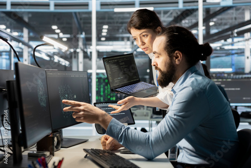 Data center manager supervising technician monitoring system performance using PC, identifying bottlenecks. Server farm teal leader overseeing worker troubleshooting glitches