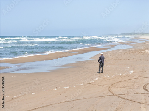 On a breezy spring day in May by the Sea of Japan, a Japanese man in his late seventies walks alone on the beach, captured from above in an aerial photo showing waves and quiet solitude.
