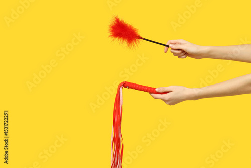 Quadro su tela Female hands with feather stick and and whip from sex shop on yellow background
