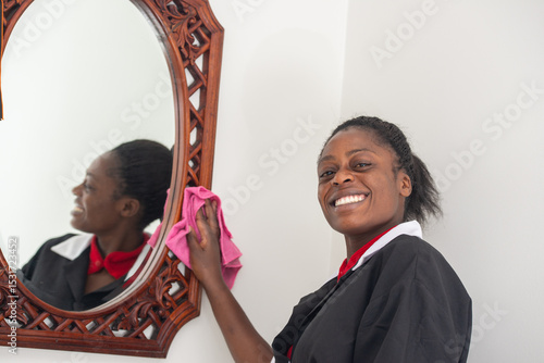 A cheerful housekeeper in uniform smiling while wiping a decorative mirror with a pink cloth.