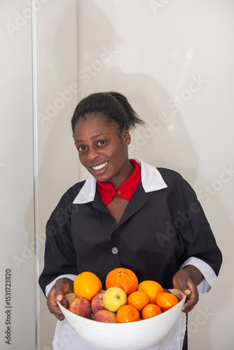 Portrait of a cheerful domestic worker in uniform holding a white bowl filled with fresh apples and citrus fruits indoors.