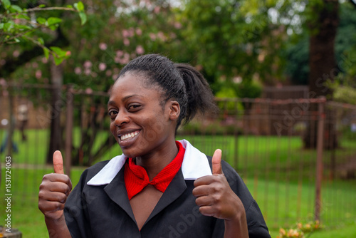Happy domestic worker in a black uniform giving thumbs up outside in a green garden with a cheerful expression.