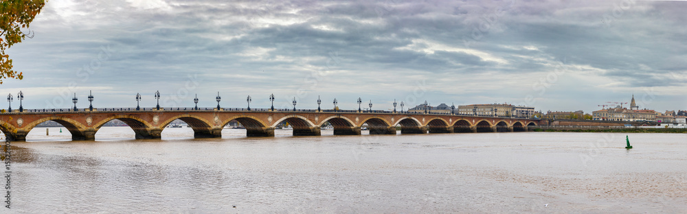Fototapeta premium Panoramic view of Pont de Pierre, a stone bridge with multiple arches and lampposts, spanning the Garonne River under a cloudy sky. Bordeaux, Gironde, Nouvelle-Aquitaine, France.