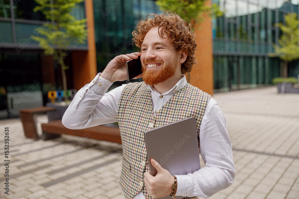 Fototapeta premium A smiling young man holds a laptop while talking on his phone in a contemporary business environment.