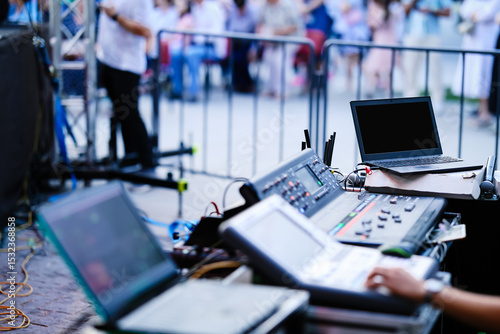 Professional audio and lighting control station at an outdoor event. Digital mixers, laptops, and control panels in use, with a blurred crowd in the background. Live event production setup.