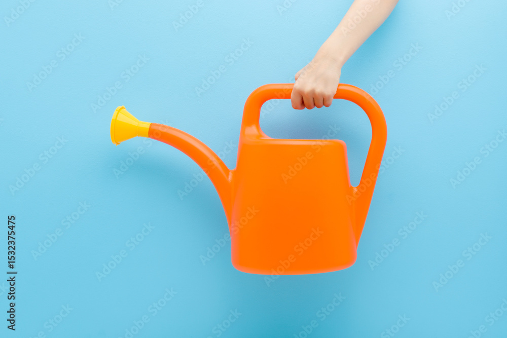 Obraz premium Little child boy hand holding and showing orange plastic can for flowers and plants watering on light blue wall background. Pastel color. Closeup. Front view.