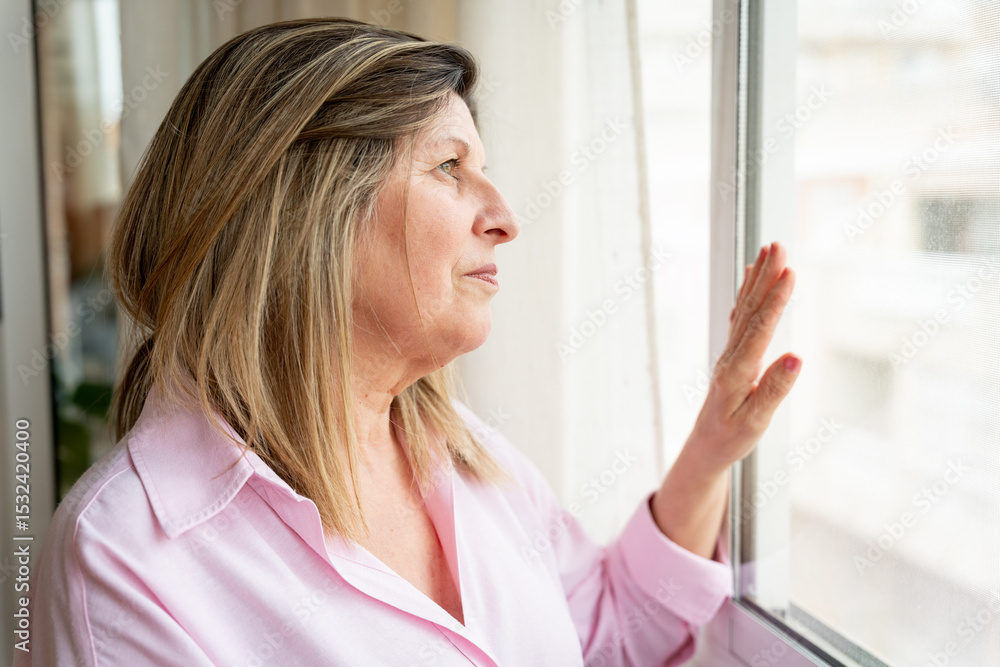 Fototapeta premium Worried senior woman resting her hand on window glass, gazing outside and yearning for brighter days ahead amidst feelings of solitude