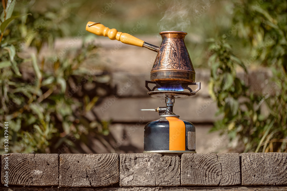 Obraz premium Fragrant coffee steams in copper engraved coffee pot on gas burner with blue flame on wooden bars. Selective focus..