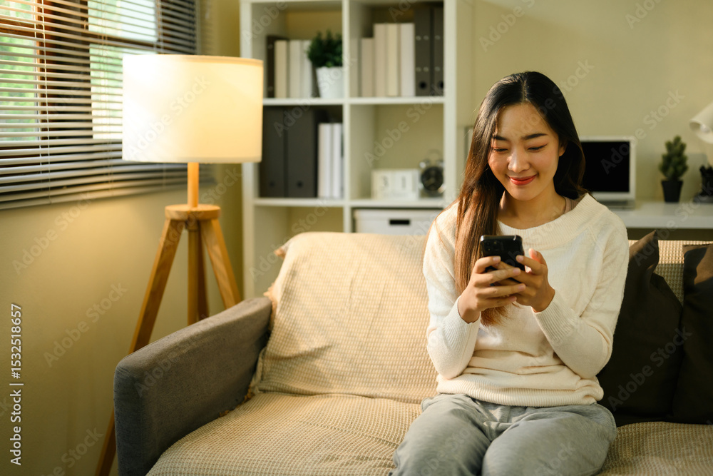 Obraz premium Young Asian Woman Relaxing at Home with Smartphone on Cozy Sofa
