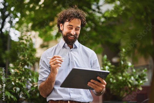 фотографія A man with curly hair and a beard is smiling as he uses a tablet in a green, bustling outdoor space
