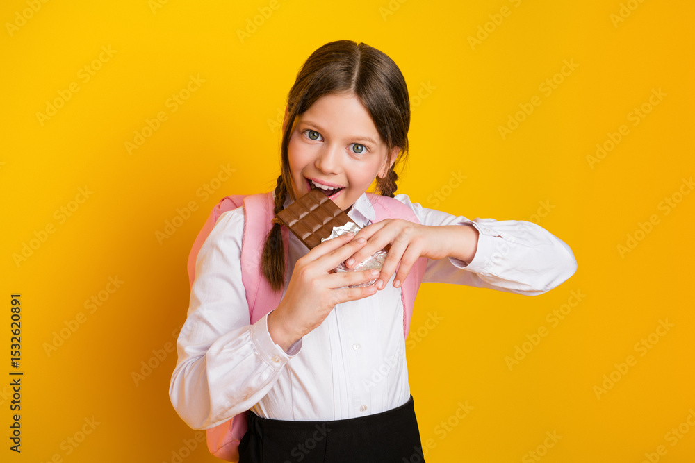 Smiling schoolgirl holding chocolate bar in hands standing against vibrant yellow background wearing uniform with braids Smiling schoolgirl holding chocolate bar in hands standing against vibrant yellow background wearing uniform with braids