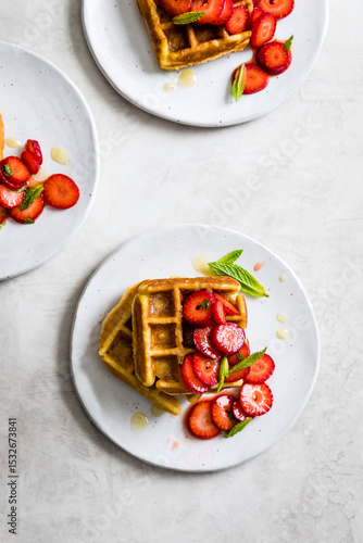 Plates of waffles topped with fresh strawberries and mint leaves on a light background.