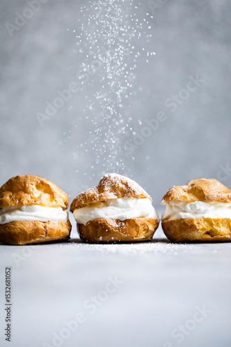 Three cream puffs being sprinkled with powdered sugar on a gray background.