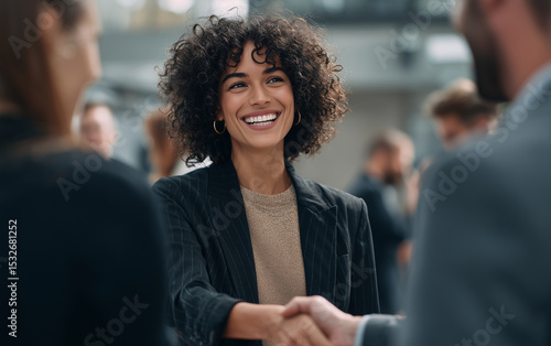  A photo of an African American woman in business attire shaking hands with another person, both smiling and looking happy as they stand amidst other people talking to eachother at the office party . 