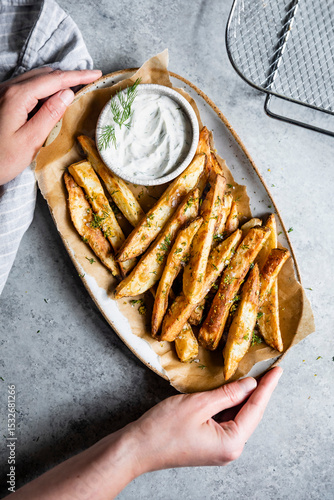 A person holding a plate of seasoned potato wedges with a bowl of dill sauce on a grey surface.