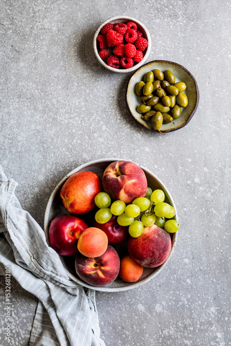 An assortment of fresh fruits including peaches, nectarines, grapes, and raspberries along with a bowl of green olives on a textured surface.
