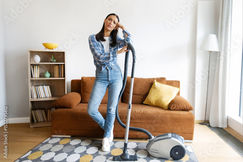 Canvas Print Tired woman with vacuum cleaner standing in modern living room, showing fatigue