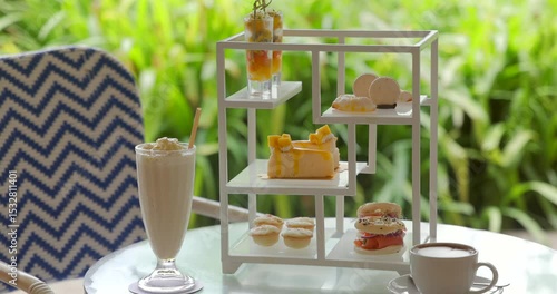 Afternoon tea set on elegant table in tropical garden, featuring refreshing milkshake, delectable desserts, macarons, and coffee. Cozy, relaxing outdoor cafe ambiance.