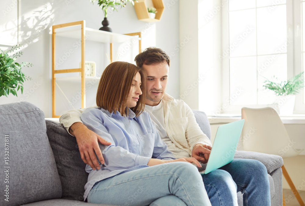 Young family, loving spouses sitting close on sofa, relaxing together at home using laptop computer, buying through e-commerce services, watching new movie online, websurfing, using new app on weekend Young family, loving spouses sitting close on sofa, relaxing together at home using laptop computer, buying through e-commerce services, watching new movie online, websurfing, using new app on weekend