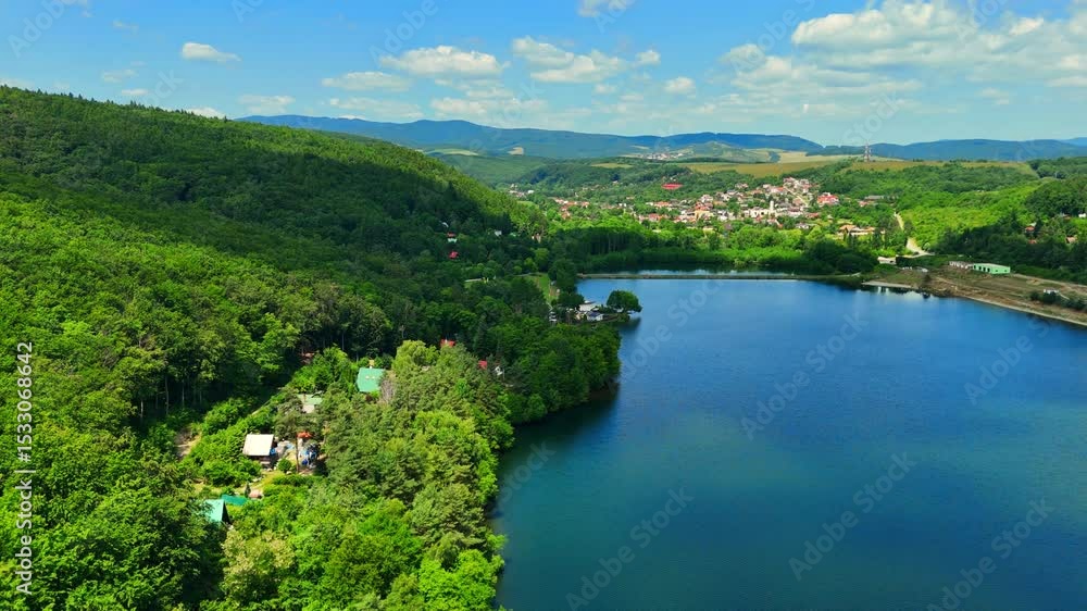 Blue calm lake among green forest in mountainous area. Countryside residential houses. Aerial view of European landscape. Slovakia. Blue calm lake among green forest in mountainous area. Countryside residential houses. Aerial view of European landscape. Slovakia.