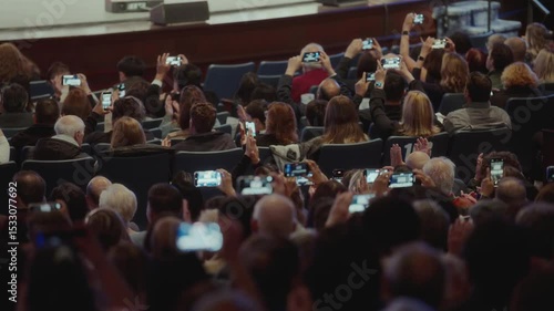 Audience at indoor event capturing performance on smartphones. People seated in rows, recording stage moments with mobile phones. Modern digital culture and media habits