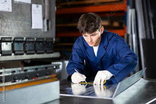 Foto Young guy, skilled worker in metal workshop, focusing on accurately marking steel sheet with tape measure, preparing workpiece for further cutting, drilling, or assembling components